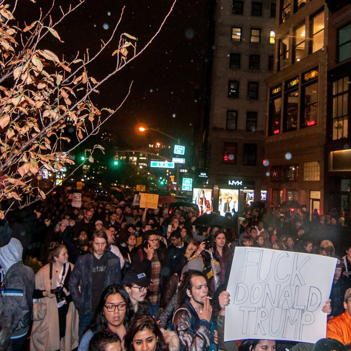 City scene at night with a large crowd of people marching through a street in the rain. In the foreground a sign is visible that reads 'Fuck Donald Trump'.
