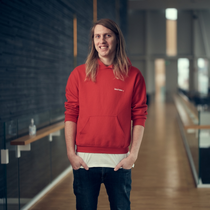 Person with long blonde hair in a bright red hoodie stands in modern-looking hotel lobby.